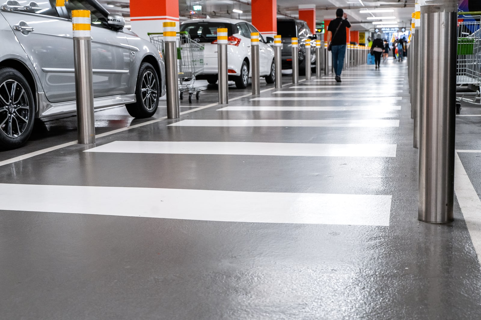 White pedestrian crossing markings on a grey internal car park floor, with bollards separating parked vehicles